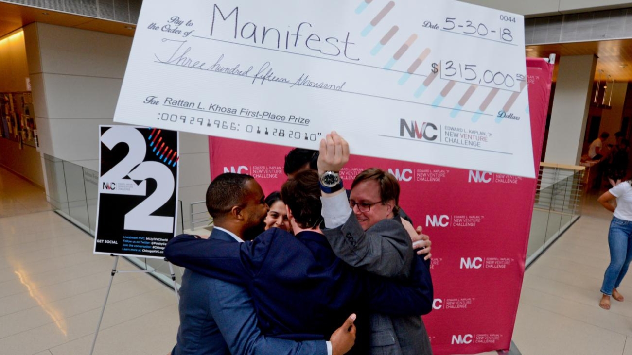 Photograph of the Manifest team hugging and holding up their poster-sized check in the amount of $315,000. Promotional material for the New Venture Challenge is in the background.
